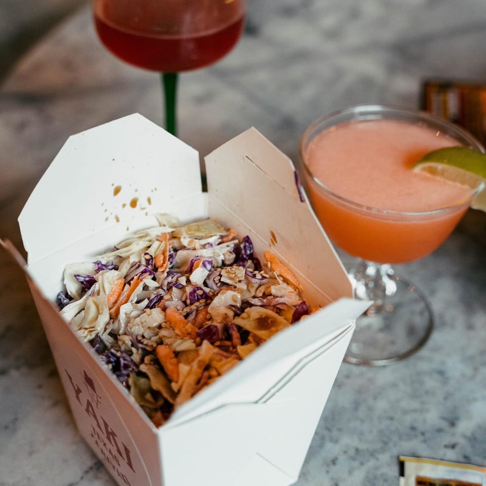 Yaki Texas Teriyaki coleslaw in a takeout box with two cocktails on a gray table.